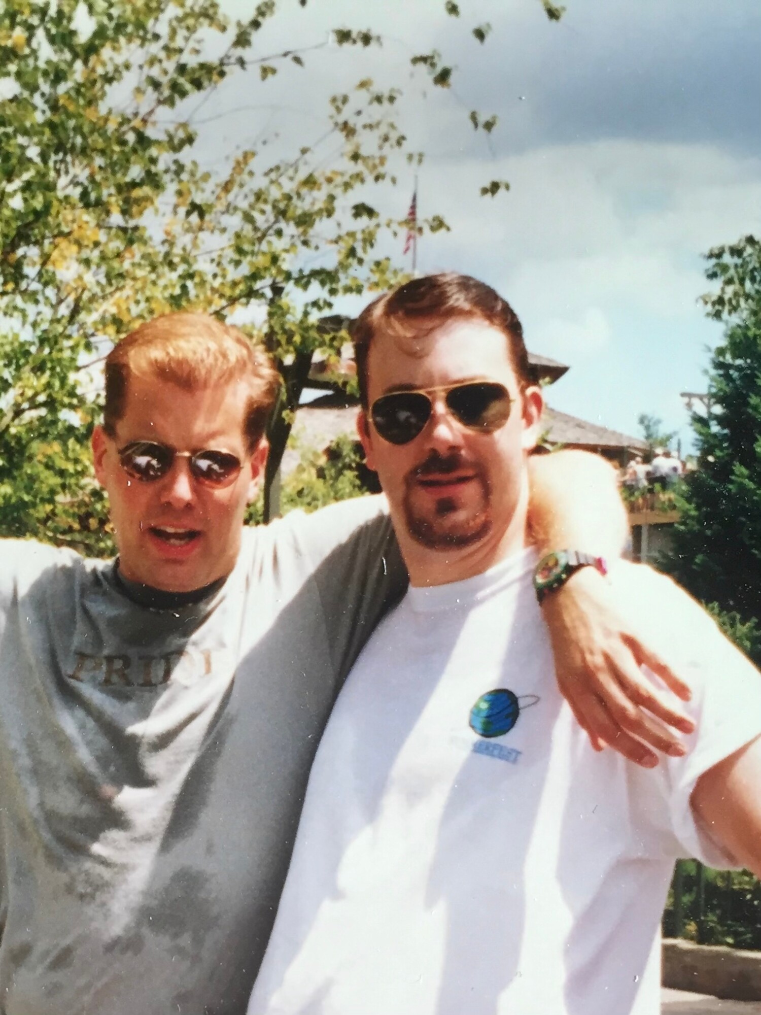 Two men look into the camera and smile with one wearing a Pride shirt and wrapping his arm around his friend